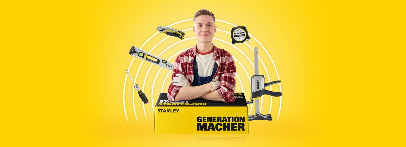 Eine Person mit unscharfem Gesicht steht hinter einer gelben STANLEY-Box mit der Aufschrift 'GENERATION MACHER' und 'STARTER-BOX', umgeben von STANLEY Handwerkzeugen wie Maßband, Wasserwaage, Cutter und Schraubendreher, vor gelbem Hintergrund.
