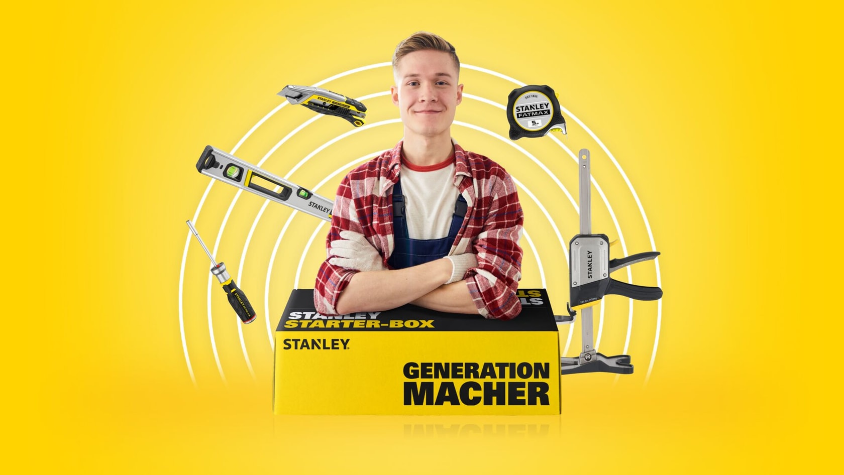 Eine Person mit verschränkten Armen steht hinter einer gelben STANLEY Starter-Box mit der Aufschrift 'GENERATION MACHER'. Verschiedene STANLEY Werkzeuge, darunter ein Maßband, eine Zwinge, eine Wasserwaage, ein Cutter und ein Schraubendreher, schweben umher. Der Hintergrund ist leuchtend gelb mit weißen Kreisen.
